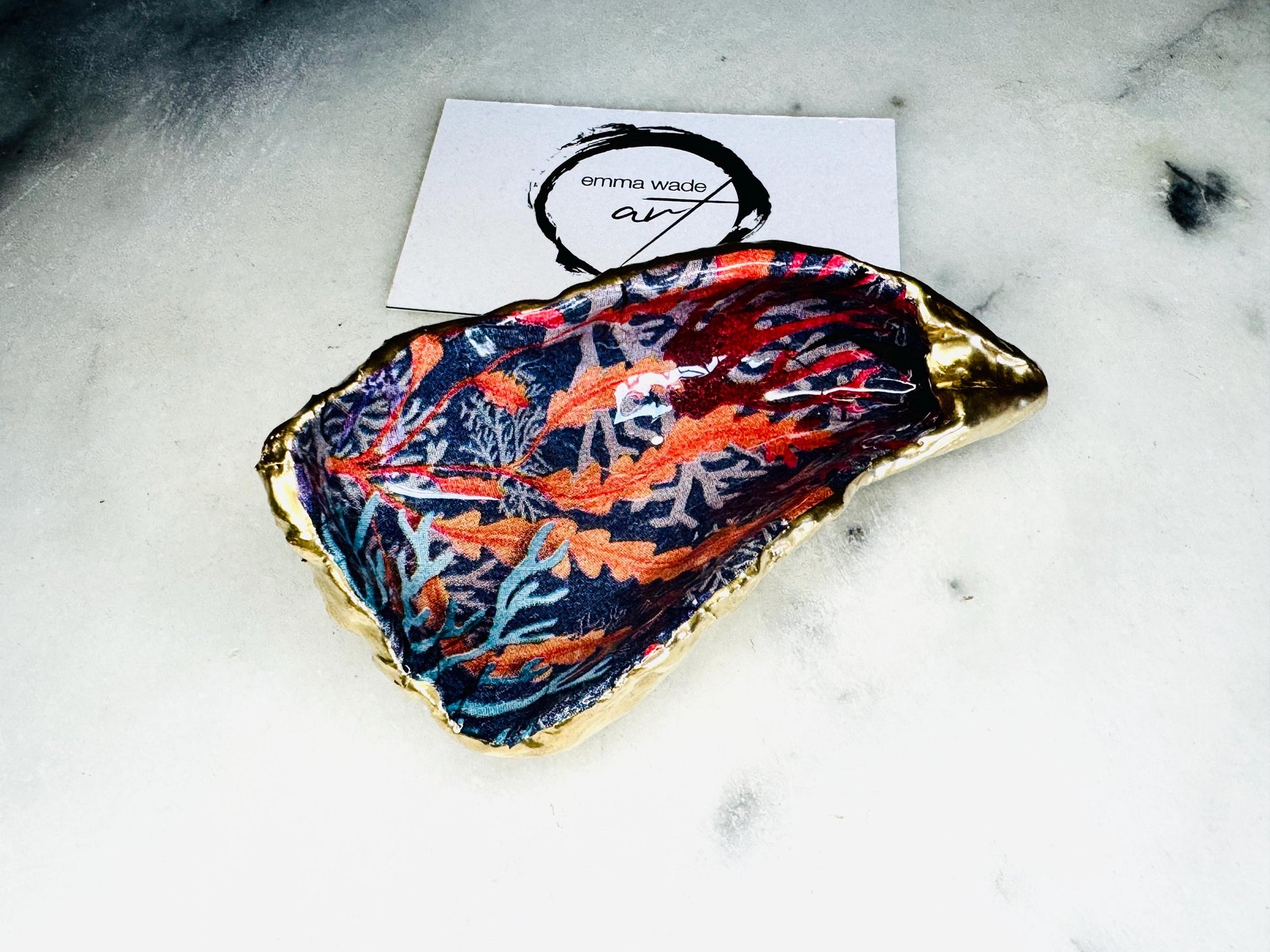 Handmade Dorset oyster shell trinket dishes featuring vibrant sea coral inspired artwork in coral red, navy and turquoise tones, finished with hand-painted gold leaf edging and glossy crystal clear resin. Coastal jewellery dishes styled on a white marble background with rich ocean-inspired botanical patterns and luxurious gilded edges.