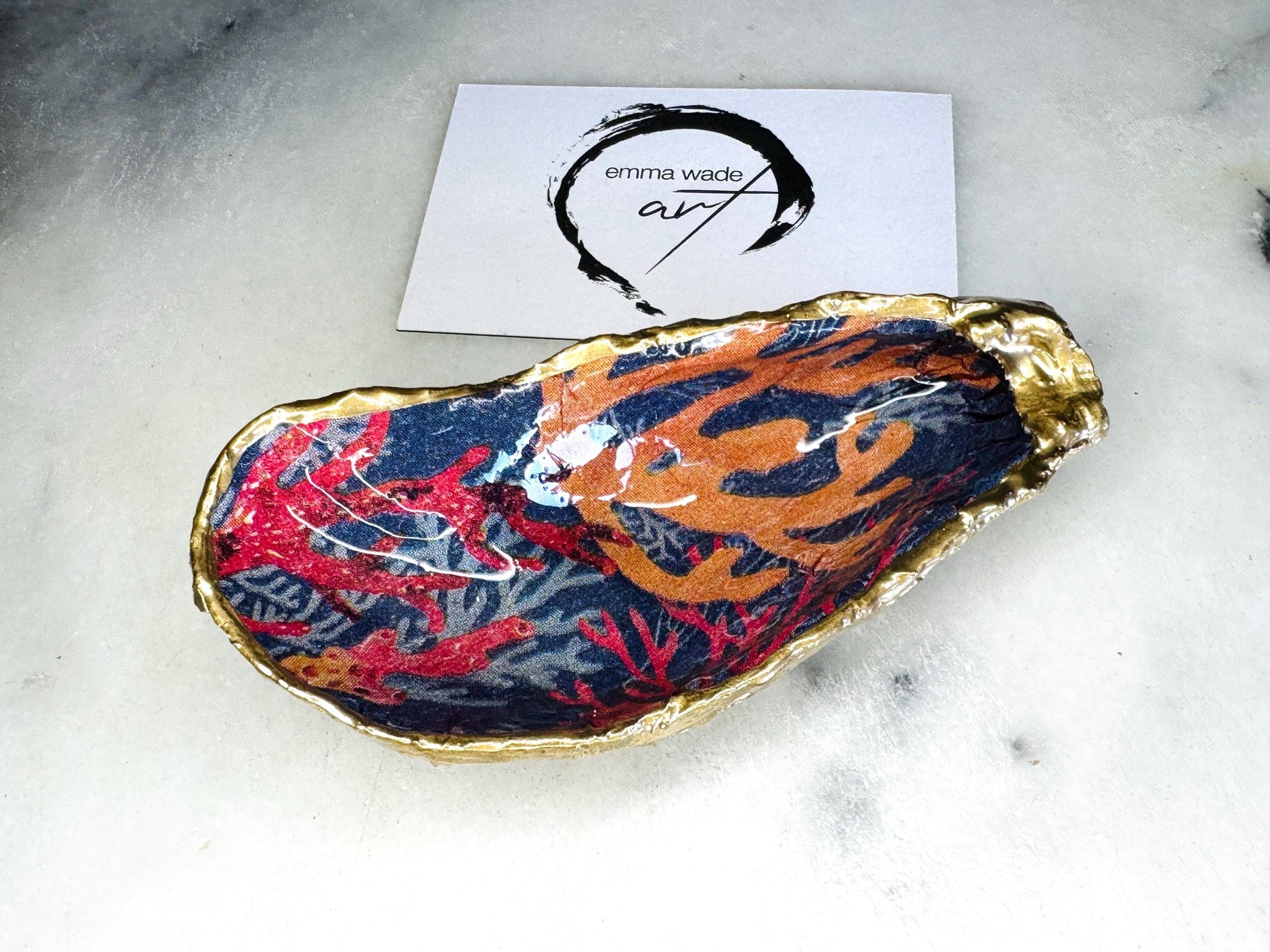 Handmade Dorset oyster shell trinket dishes featuring vibrant sea coral inspired artwork in coral red, navy and turquoise tones, finished with hand-painted gold leaf edging and glossy crystal clear resin. Coastal jewellery dishes styled on a white marble background with rich ocean-inspired botanical patterns and luxurious gilded edges.