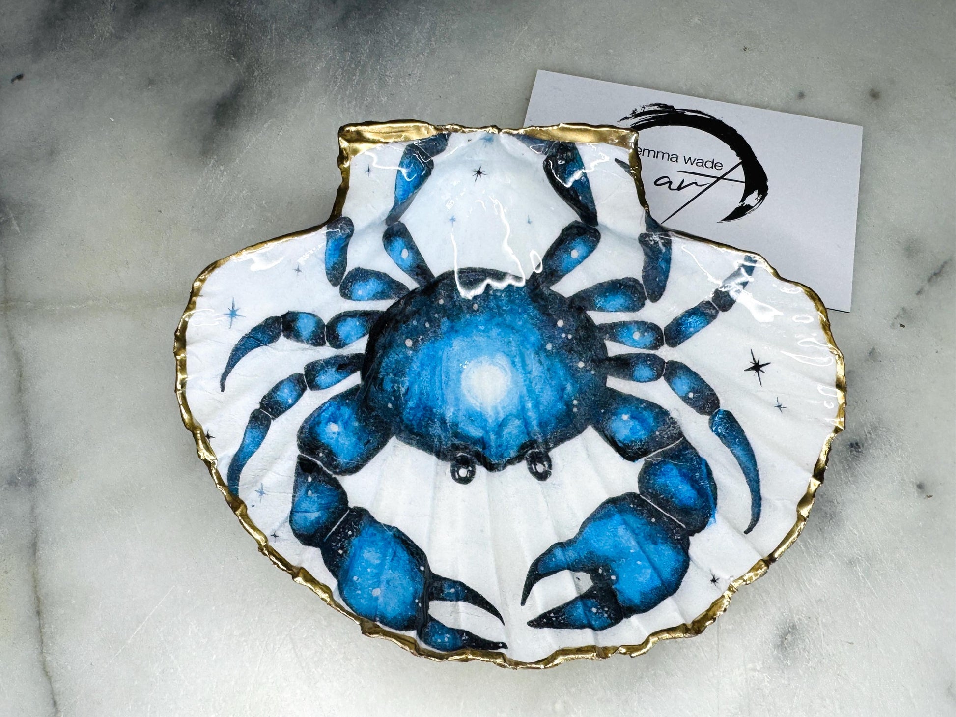 Scallop shell trinket dish with a hand drawn and hand painted blue crab design from original watercolour artwork. The artwork is applied as a decoupage print, then enhanced with painted detailing for a unique finish. Each shell is edged with liquid gold leaf and sealed with glossy resin. A handmade shell ring dish, jewellery organiser, or trinket tray for rings, earrings, or cufflinks. Perfect coastal decor or gift for weddings, anniversaries, birthdays, or Christmas.