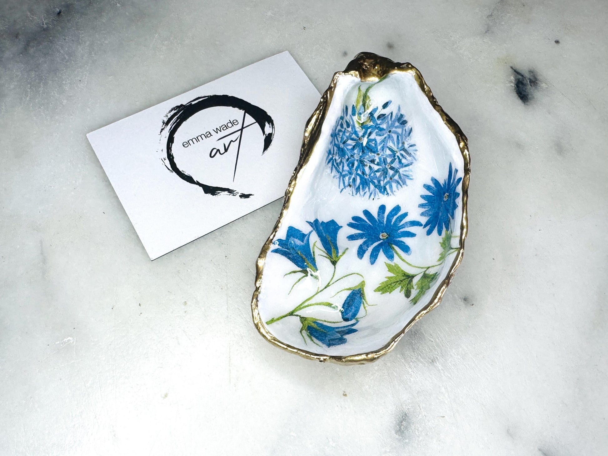 Handmade oyster shell jewellery dish with blue spring flower design, finished with gold leaf edging and glossy resin. Each Dorset oyster shell is unique, making a one-of-a-kind jewellery tray, ring holder, or trinket dish for rings, earrings, cufflinks, or keepsakes. A beautiful shell jewellery catcher and organiser with floral detail, perfect as a handmade gift for weddings, anniversaries, birthdays, bridesmaids, or Christmas. Elegant coastal home decor with botanical charm.