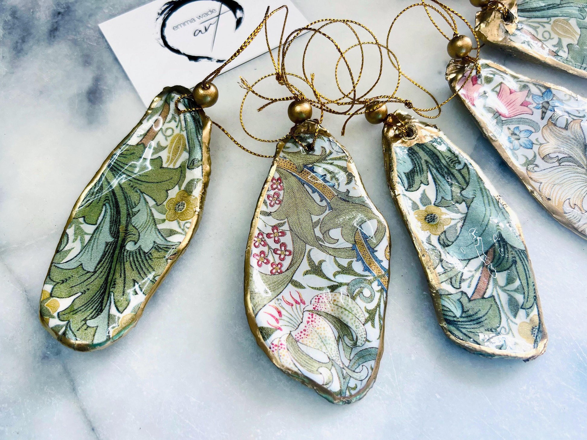 Handmade Dorset oyster shell hanging ornaments featuring William Morris inspired Golden Lily and Granville floral designs in soft green, blush pink and gold tones. Natural oyster shells finished with liquid gold leaf edging and sealed in glossy resin. Decorative hanging ornaments with gold cord and bead detail, suitable for luxury Christmas décor, coastal interiors, cottagecore styling or elegant year-round home decoration.