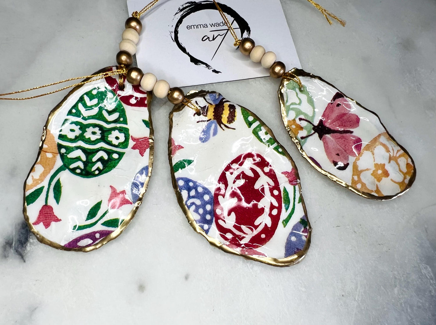 Handmade Dorset oyster shell ornaments featuring Emma Bridgewater bee, butterfly, and egg designs. Each shell has a natural pearl finish, gilded with liquid gold leaf on the rim and back, and sealed with glossy resin for shine and durability. Available individually or as a set of 3, with pre-drilled holes, gold hanging string, and decorative beads. Unique festive decorations for Christmas trees, seasonal home décor, or handmade coastal-inspired gifts for weddings, birthdays, or anniversaries.