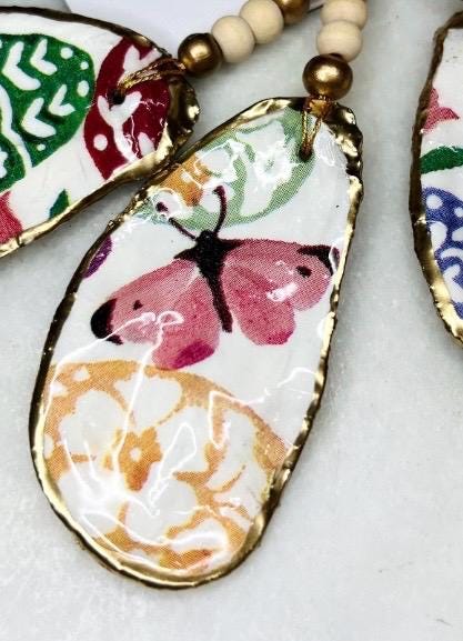 Handmade Dorset oyster shell ornaments featuring Emma Bridgewater bee, butterfly, and egg designs. Each shell has a natural pearl finish, gilded with liquid gold leaf on the rim and back, and sealed with glossy resin for shine and durability. Available individually or as a set of 3, with pre-drilled holes, gold hanging string, and decorative beads. Unique festive decorations for Christmas trees, seasonal home décor, or handmade coastal-inspired gifts for weddings, birthdays, or anniversaries.