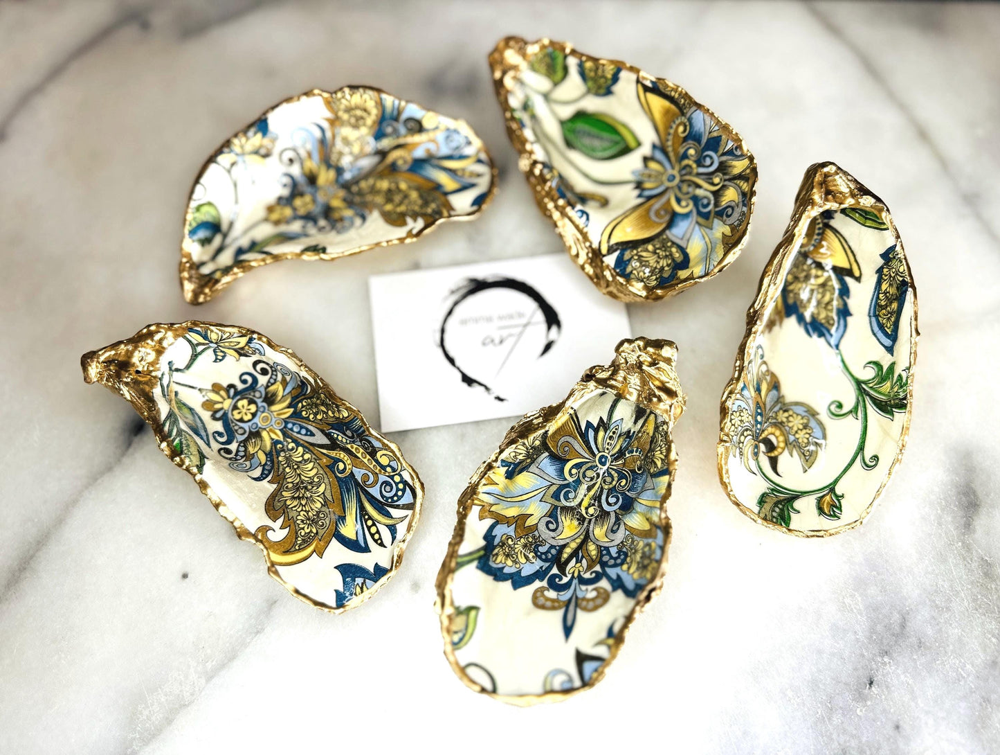 Handmade Dorset oyster shell trinket dishes decorated with ornate blue, green and gold baroque floral patterns inspired by antique European porcelain and vintage decorative art. Natural oyster shells are hand gilded with liquid gold leaf edging and sealed with glossy crystal clear resin. Luxury coastal jewelry holders and vanity dishes photographed on marble background with elegant decorative styling.