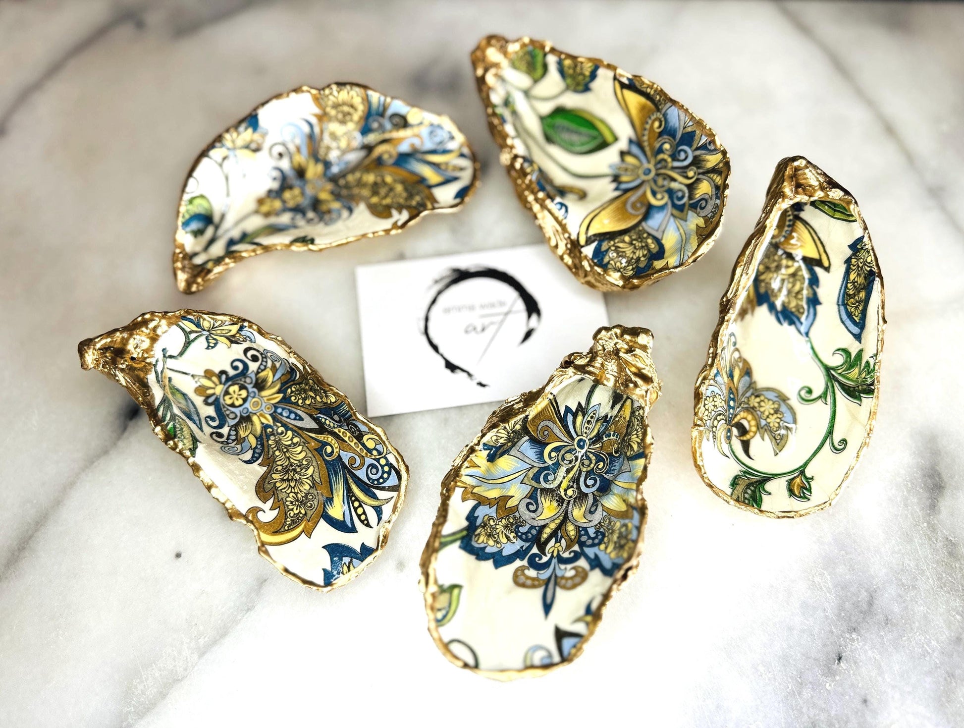 Handmade Dorset oyster shell trinket dishes decorated with ornate blue, green and gold baroque floral patterns inspired by antique European porcelain and vintage decorative art. Natural oyster shells are hand gilded with liquid gold leaf edging and sealed with glossy crystal clear resin. Luxury coastal jewelry holders and vanity dishes photographed on marble background with elegant decorative styling.