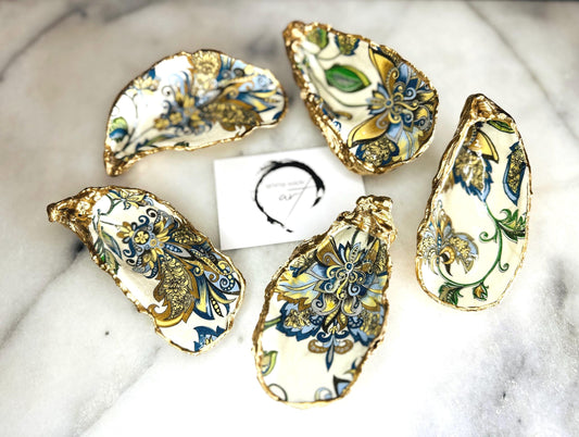 Handmade Dorset oyster shell trinket dishes decorated with ornate blue, green and gold baroque floral patterns inspired by antique European porcelain and vintage decorative art. Natural oyster shells are hand gilded with liquid gold leaf edging and sealed with glossy crystal clear resin. Luxury coastal jewelry holders and vanity dishes photographed on marble background with elegant decorative styling.
