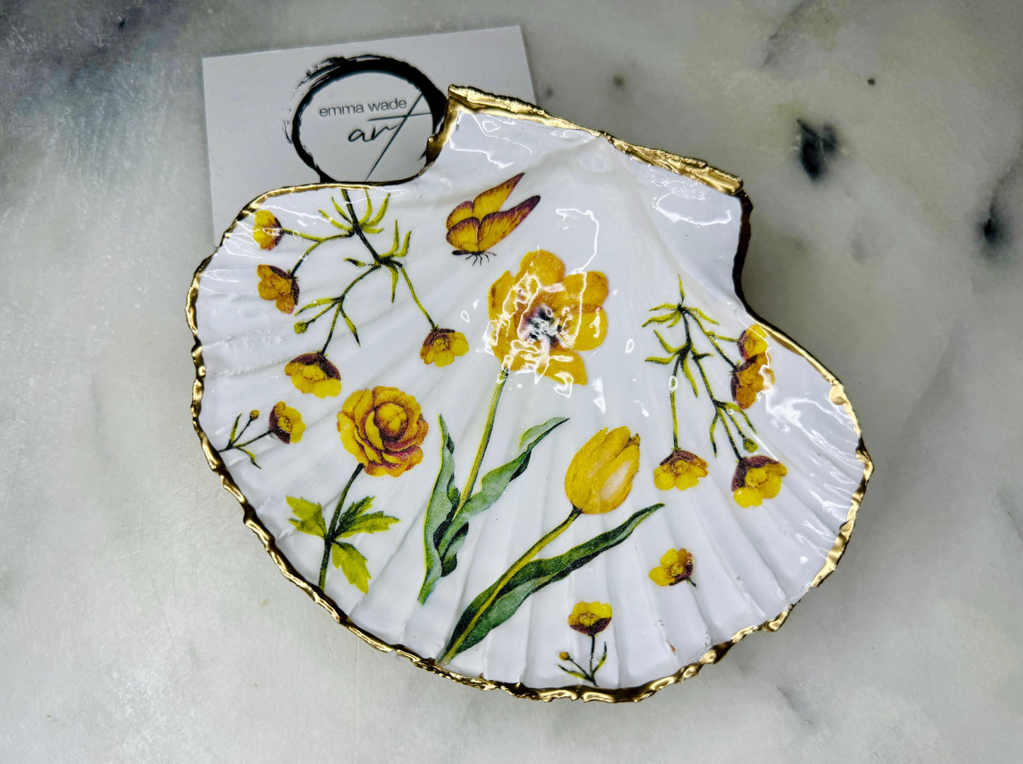 Hand finished scallop shell trinket dish featuring yellow spring flower artwork with a hand gilded gold leaf rim, sealed in glossy clear resin for durability and shine. Coastal inspired shell jewellery dish photographed on a light surface, ideal for rings, earrings and small keepsakes. Decorative floral shell dish by EmmaWadeART inspired by the Dorset coastline, perfect as a gift or elegant home décor accent.