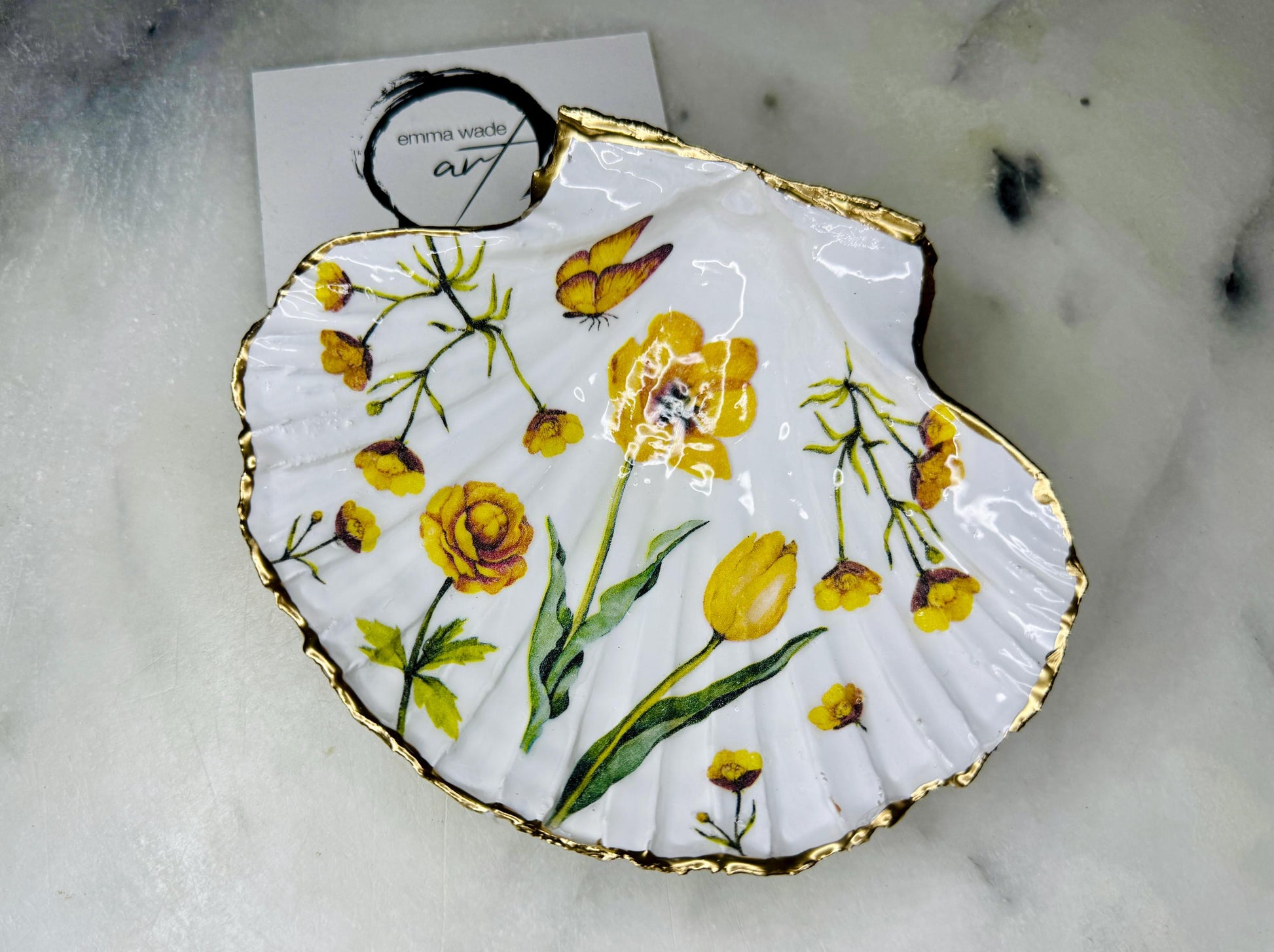 Hand finished scallop shell trinket dish featuring yellow spring flower artwork with a hand gilded gold leaf rim, sealed in glossy clear resin for durability and shine. Coastal inspired shell jewellery dish photographed on a light surface, ideal for rings, earrings and small keepsakes. Decorative floral shell dish by EmmaWadeART inspired by the Dorset coastline, perfect as a gift or elegant home décor accent.