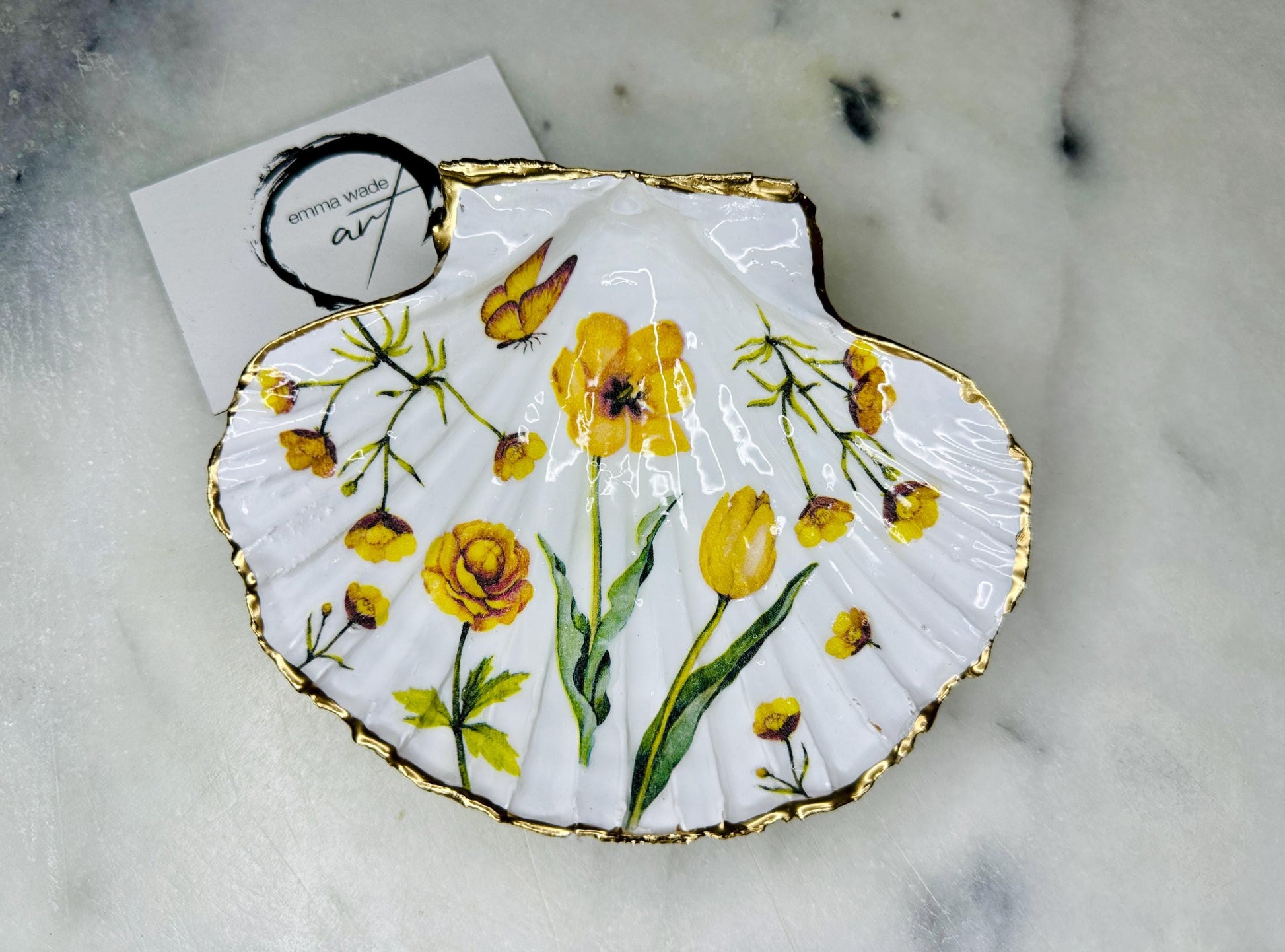 Hand finished scallop shell trinket dish featuring yellow spring flower artwork with a hand gilded gold leaf rim, sealed in glossy clear resin for durability and shine. Coastal inspired shell jewellery dish photographed on a light surface, ideal for rings, earrings and small keepsakes. Decorative floral shell dish by EmmaWadeART inspired by the Dorset coastline, perfect as a gift or elegant home décor accent.