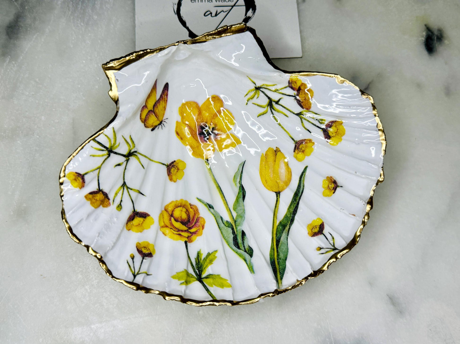 Hand finished scallop shell trinket dish featuring yellow spring flower artwork with a hand gilded gold leaf rim, sealed in glossy clear resin for durability and shine. Coastal inspired shell jewellery dish photographed on a light surface, ideal for rings, earrings and small keepsakes. Decorative floral shell dish by EmmaWadeART inspired by the Dorset coastline, perfect as a gift or elegant home décor accent.