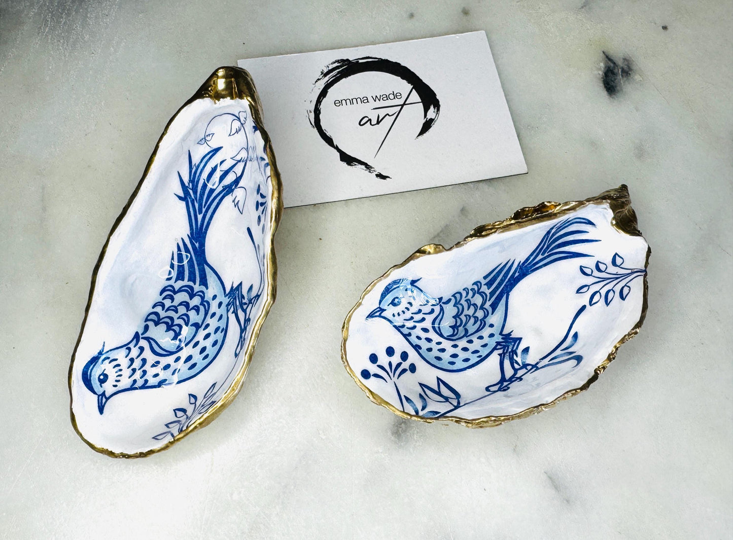 Handmade Dorset oyster shell trinket dish with blue porcelain-style bird design, gold leaf gilded edges and glossy resin finish. Coastal jewellery holder and ring dish photographed on a light surface, showing the natural shell shape and pearly interior. Unique sustainably sourced shell, sold individually as decorative coastal home décor or a thoughtful gift.