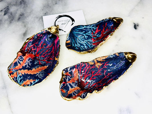 Handmade Dorset oyster shell trinket dishes featuring vibrant sea coral inspired artwork in coral red, navy and turquoise tones, finished with hand-painted gold leaf edging and glossy crystal clear resin. Coastal jewellery dishes styled on a white marble background with rich ocean-inspired botanical patterns and luxurious gilded edges.