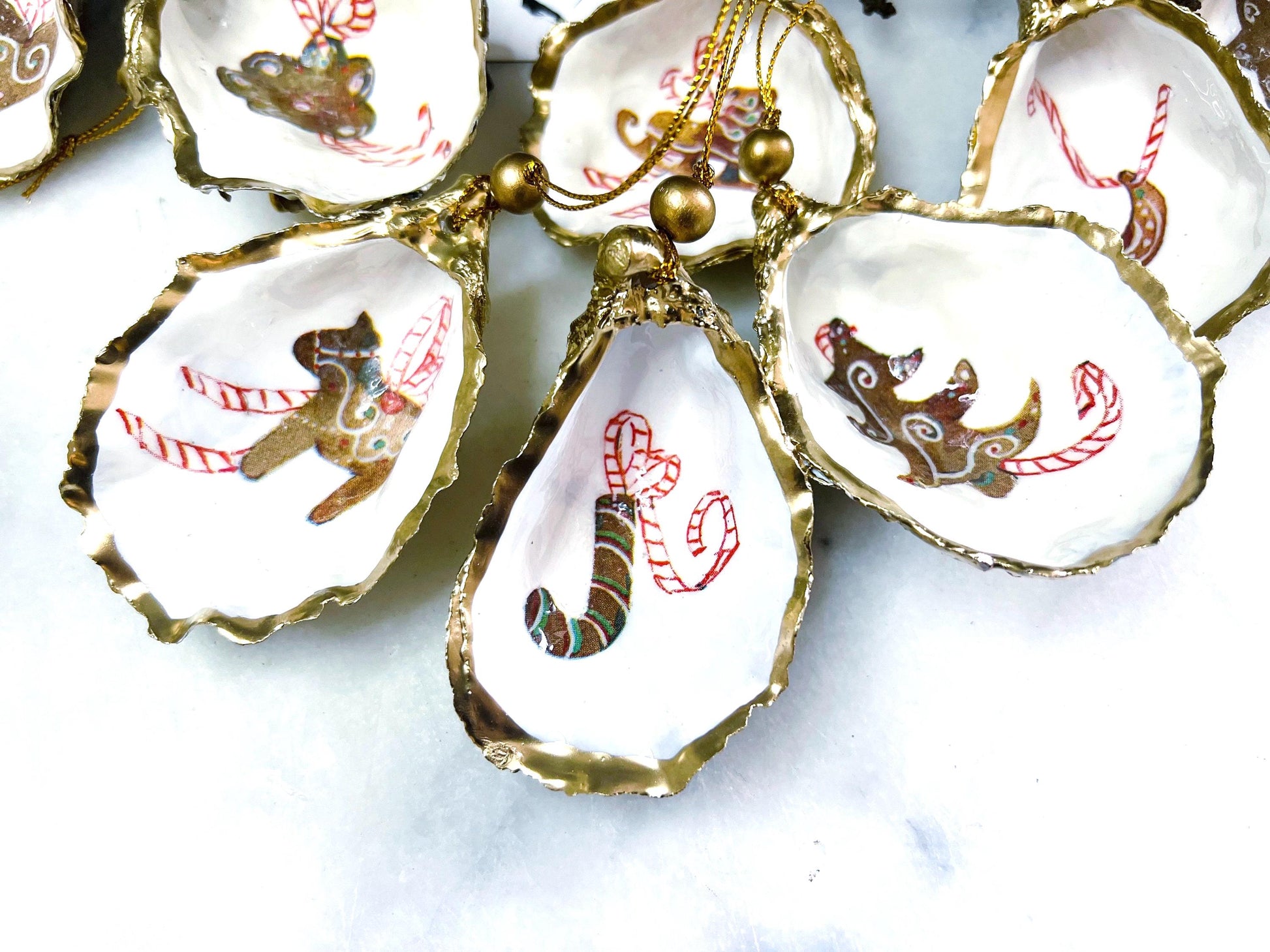 Handcrafted Dorset oyster shell Christmas ornaments featuring gingerbread-inspired festive motifs including candy canes, stars, moons, hearts, and skates. Gold leaf edged coastal Christmas baubles with glossy resin finish and pearl shell interiors displayed as luxury handmade holiday decorations.