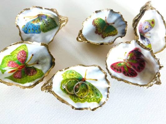 Handmade Dorset oyster shell ring dishes featuring colourful butterfly designs in green, blue and pink tones. Decorative oyster shell trinket holders finished with hand-applied liquid gold leaf edging and glossy resin. Nature inspired jewellery dishes with natural pearlescent shell interiors, styled as coastal décor, ring holders and keepsake gifts.