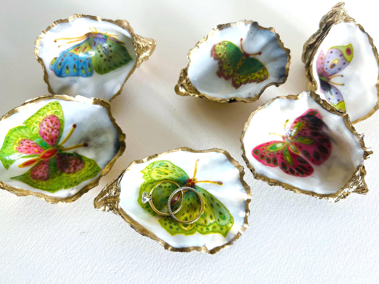 Handmade Dorset oyster shell ring dishes featuring colourful butterfly designs in green, blue and pink tones. Decorative oyster shell trinket holders finished with hand-applied liquid gold leaf edging and glossy resin. Nature inspired jewellery dishes with natural pearlescent shell interiors, styled as coastal décor, ring holders and keepsake gifts.