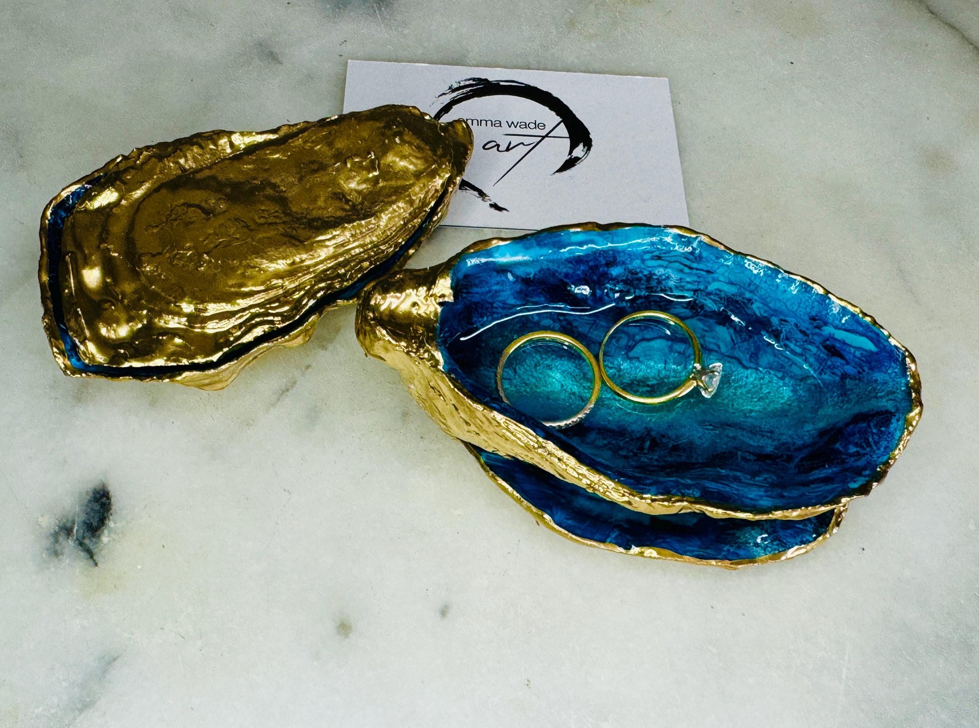 Hand-painted Dorset oyster shell ring box featuring deep blue ocean-inspired resin artwork with sapphire and turquoise wave details, matching gold leaf lid and glossy crystal-clear finish. Luxury coastal proposal ring holder and wedding keepsake box styled with gold rings, inspired by sea caves, tidal pools and the Dorset coastline. Handmade oyster shell jewellery box with hand gilded exterior and unique ocean cove design for engagements, weddings and anniversaries.