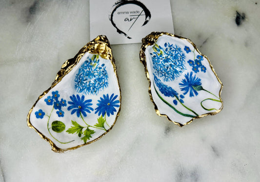 Handmade Dorset oyster shell trinket dish featuring delicate blue floral artwork including bluebells and forget-me-nots, finished with hand-applied gold leaf edging and glossy resin. Coastal-inspired jewellery dish and ring holder available in multiple sizes including XL oyster shells. Elegant handmade home décor gift ideal for birthdays, bridesmaids, Mother’s Day, weddings, and coastal interiors.