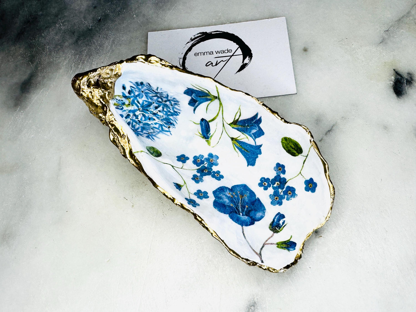 Handmade Dorset oyster shell trinket dish featuring delicate blue floral artwork including bluebells and forget-me-nots, finished with hand-applied gold leaf edging and glossy resin. Coastal-inspired jewellery dish and ring holder available in multiple sizes including XL oyster shells. Elegant handmade home décor gift ideal for birthdays, bridesmaids, Mother’s Day, weddings, and coastal interiors.