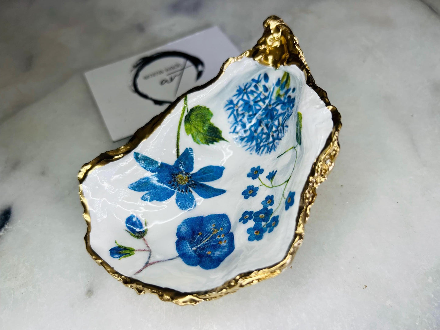 Handmade Dorset oyster shell trinket dish featuring delicate blue floral artwork including bluebells and forget-me-nots, finished with hand-applied gold leaf edging and glossy resin. Coastal-inspired jewellery dish and ring holder available in multiple sizes including XL oyster shells. Elegant handmade home décor gift ideal for birthdays, bridesmaids, Mother’s Day, weddings, and coastal interiors.