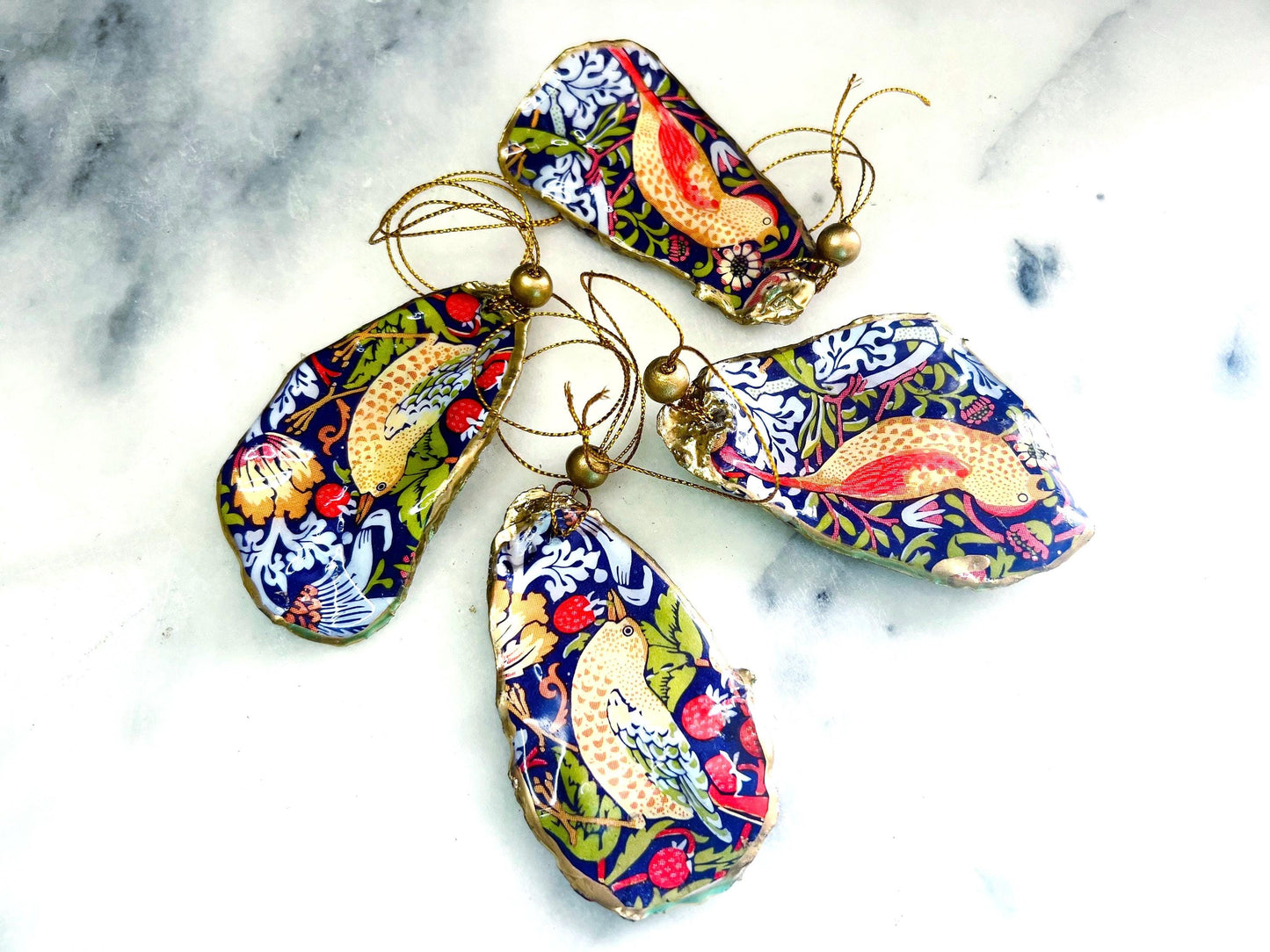 Handmade flat oyster shell ornaments decorated with William Morris Red Strawberry Thief design. Each Dorset shell has a natural pearl finish, gilded with liquid gold leaf on the rim and back, and sealed with glossy resin for durability and shine. Pre-drilled with gold string and bead for easy hanging. Available individually, as a set of 3, or set of 6. Unique coastal-inspired decorations, perfect as Christmas tree ornaments, wedding favours, bridesmaid gifts, or handmade William Morris presents.