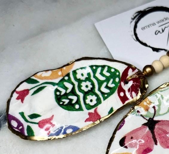 Handmade Dorset oyster shell ornaments featuring Emma Bridgewater bee, butterfly, and egg designs. Each shell has a natural pearl finish, gilded with liquid gold leaf on the rim and back, and sealed with glossy resin for shine and durability. Available individually or as a set of 3, with pre-drilled holes, gold hanging string, and decorative beads. Unique festive decorations for Christmas trees, seasonal home décor, or handmade coastal-inspired gifts for weddings, birthdays, or anniversaries.