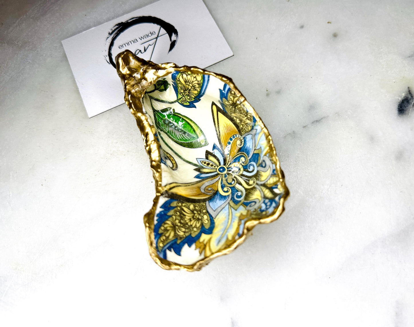 Handmade Dorset oyster shell trinket dishes decorated with ornate blue, green and gold baroque floral patterns inspired by antique European porcelain and vintage decorative art. Natural oyster shells are hand gilded with liquid gold leaf edging and sealed with glossy crystal clear resin. Luxury coastal jewelry holders and vanity dishes photographed on marble background with elegant decorative styling.