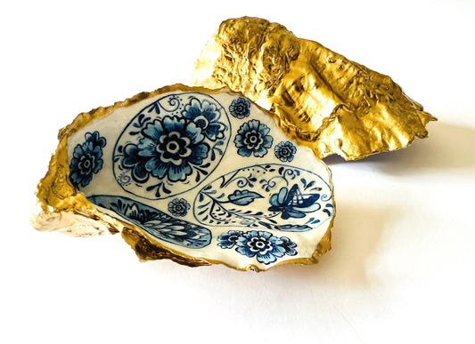 Handmade Dorset oyster shell trinket dish featuring a vintage blue and white decorative egg design with intricate floral porcelain style detailing. Oyster shell is hand gilded with liquid gold leaf edging and sealed in glossy crystal clear resin. Elegant coastal jewelry holder, ring dish, trinket tray, or decorative keepsake inspired by antique ceramics and vintage decor styling. Luxury handmade shell art photographed on a marble background.