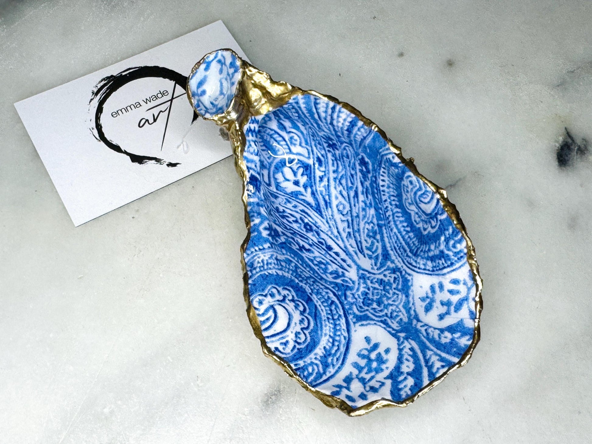 Handmade Dorset oyster shell trinket dish featuring an elegant blue and white paisley design, hand-gilded with liquid gold leaf and finished in glossy resin. Coastal shell jewellery holder available in multiple sizes from small to XL, ideal as a ring dish, trinket tray, vanity decor, or luxury coastal gift.