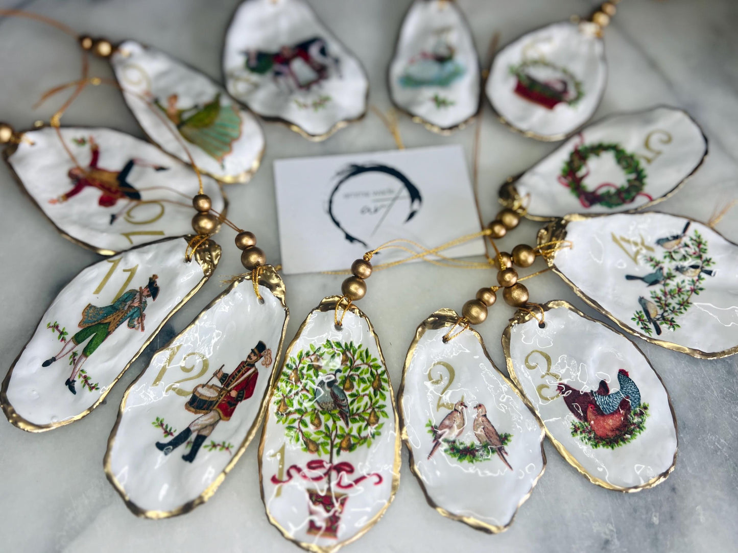 Handmade Twelve Days of Christmas oyster shell ornament set featuring all twelve traditional carol illustrations, finished with gold leaf edging, glossy resin and gold hanging cords. Coastal heirloom Christmas decorations handcrafted from Dorset oyster shells.