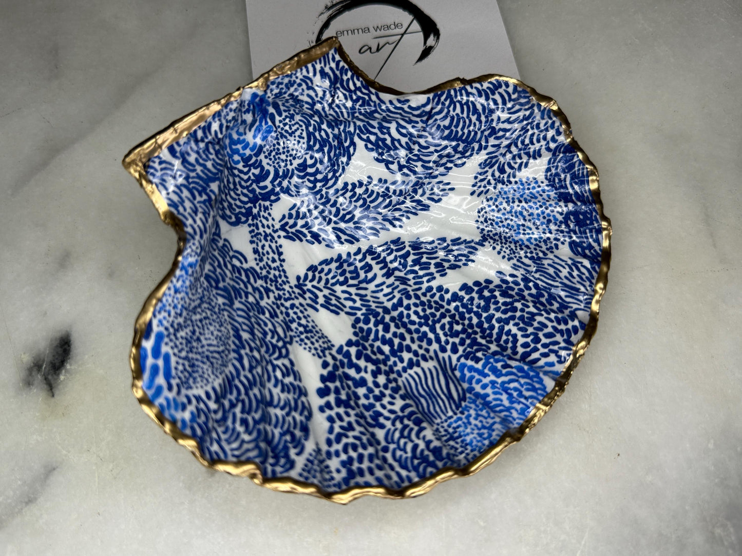 Marimekko inspired blue floral scallop shell trinket dish with hand-applied gold leaf edging and glossy resin finish. Natural Dorset shell jewellery holder featuring bold Scandinavian style botanical pattern. Decorative coastal ring dish styled on a marble surface, ideal as a bedside trinket bowl, host gift or luxury handmade home accent.
