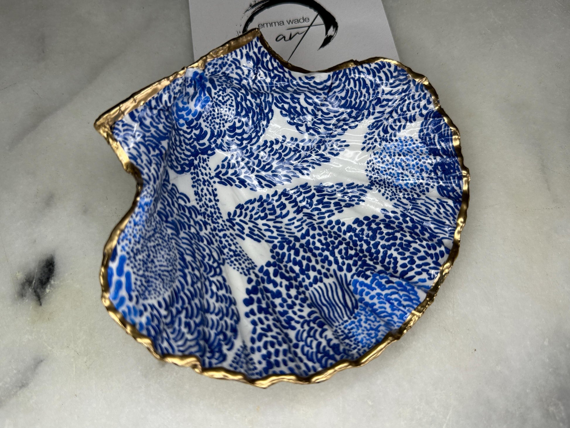 Marimekko inspired blue floral scallop shell trinket dish with hand-applied gold leaf edging and glossy resin finish. Natural Dorset shell jewellery holder featuring bold Scandinavian style botanical pattern. Decorative coastal ring dish styled on a marble surface, ideal as a bedside trinket bowl, host gift or luxury handmade home accent.