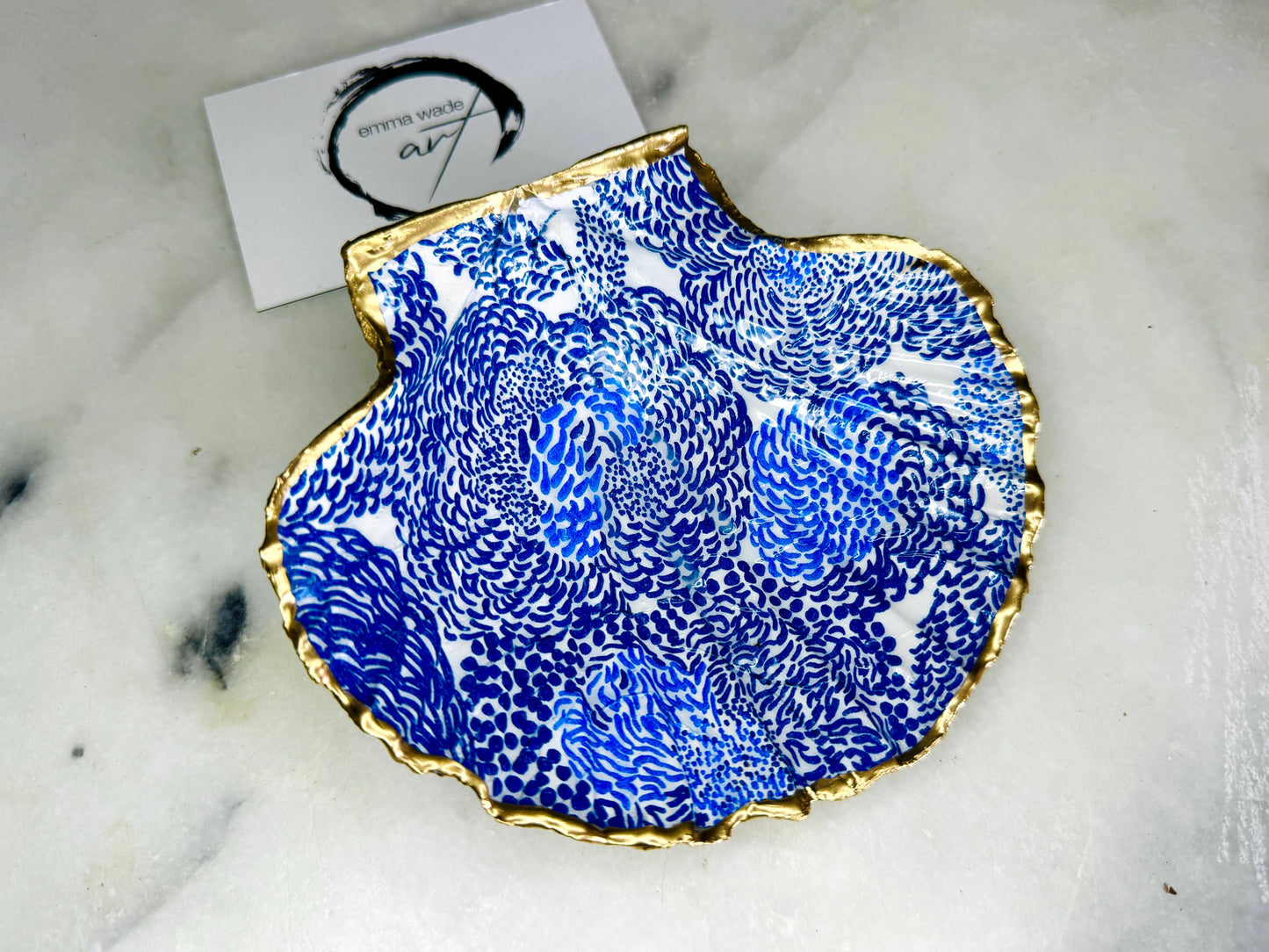 Marimekko inspired blue floral scallop shell trinket dish with hand-applied gold leaf edging and glossy resin finish. Natural Dorset shell jewellery holder featuring bold Scandinavian style botanical pattern. Decorative coastal ring dish styled on a marble surface, ideal as a bedside trinket bowl, host gift or luxury handmade home accent.
