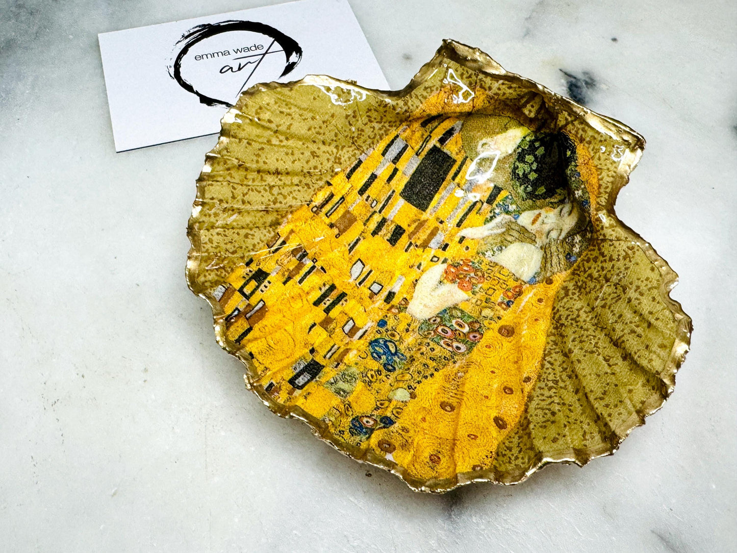 Handmade scallop shell trinket dish inspired by Gustav Klimt’s “The Kiss”, finished with gold leaf and sealed with glossy resin. Coastal Dorset shell jewellery dish perfect for rings, earrings and small treasures. Unique decorative shell bowl with golden artwork design, photographed on a light background showing detailed gilded surface and natural shell shape.