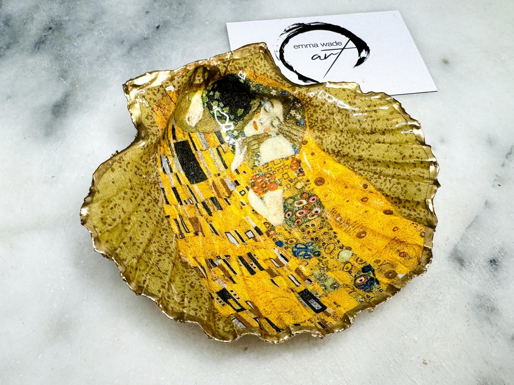 Handmade scallop shell trinket dish inspired by Gustav Klimt’s “The Kiss”, finished with gold leaf and sealed with glossy resin. Coastal Dorset shell jewellery dish perfect for rings, earrings and small treasures. Unique decorative shell bowl with golden artwork design, photographed on a light background showing detailed gilded surface and natural shell shape.