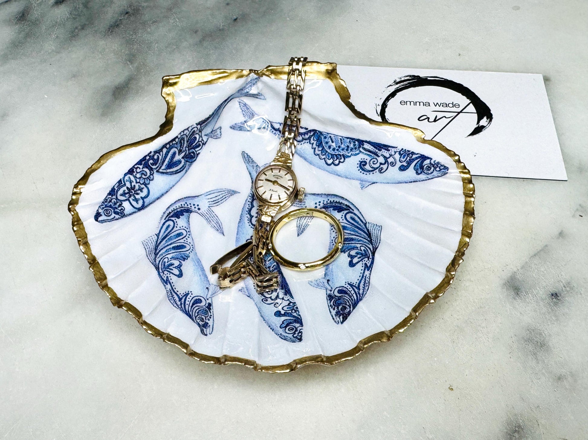 Handmade Dorset scallop shell trinket dishes featuring blue and white paisley fish illustrations with hand-painted gold leaf edging and glossy crystal clear resin. Coastal Scandinavian inspired jewellery dishes styled on a soft neutral background with elegant nautical fish artwork and luxurious gilded detailing.