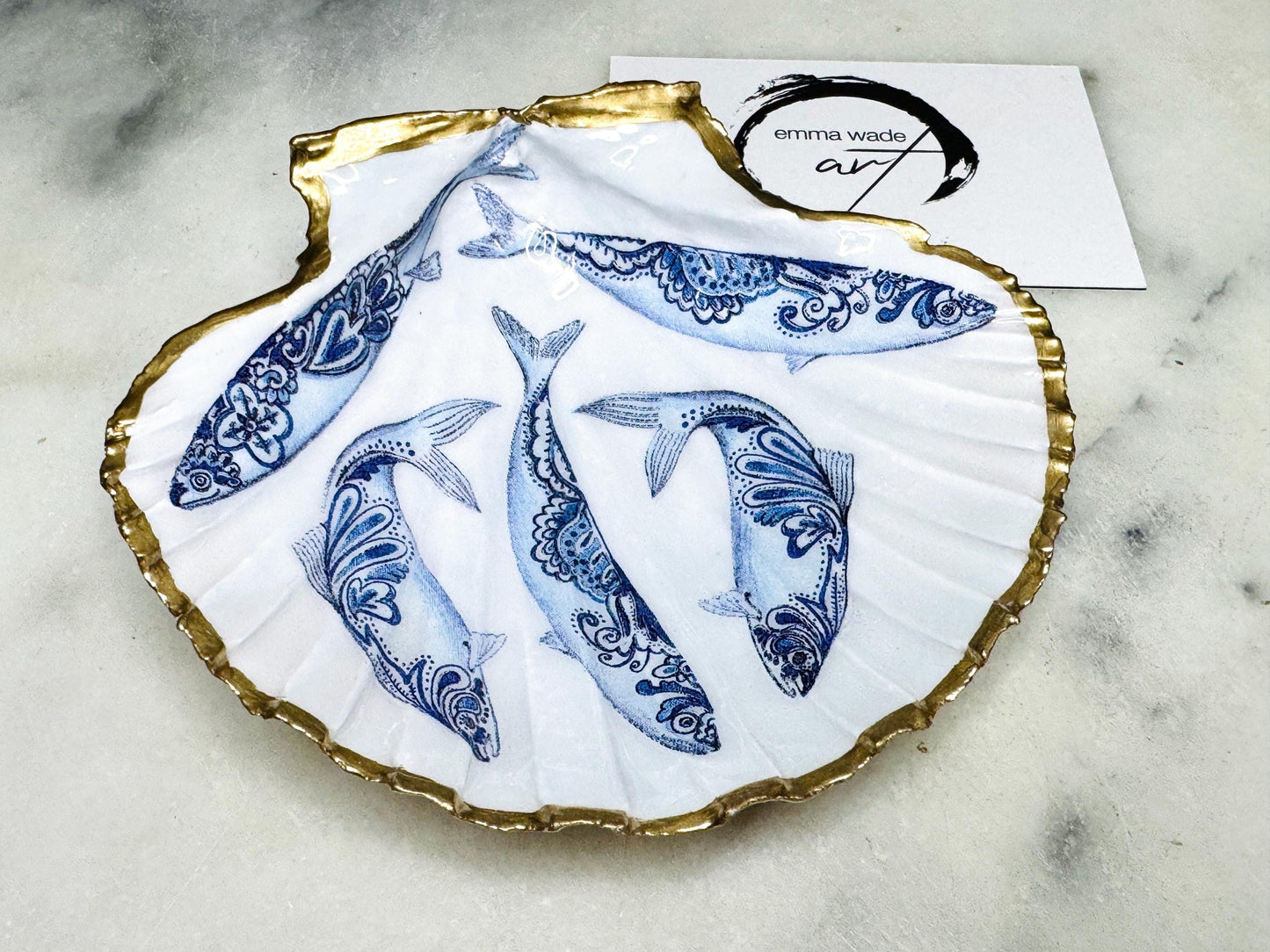 Handmade Dorset scallop shell trinket dishes featuring blue and white paisley fish illustrations with hand-painted gold leaf edging and glossy crystal clear resin. Coastal Scandinavian inspired jewellery dishes styled on a soft neutral background with elegant nautical fish artwork and luxurious gilded detailing.