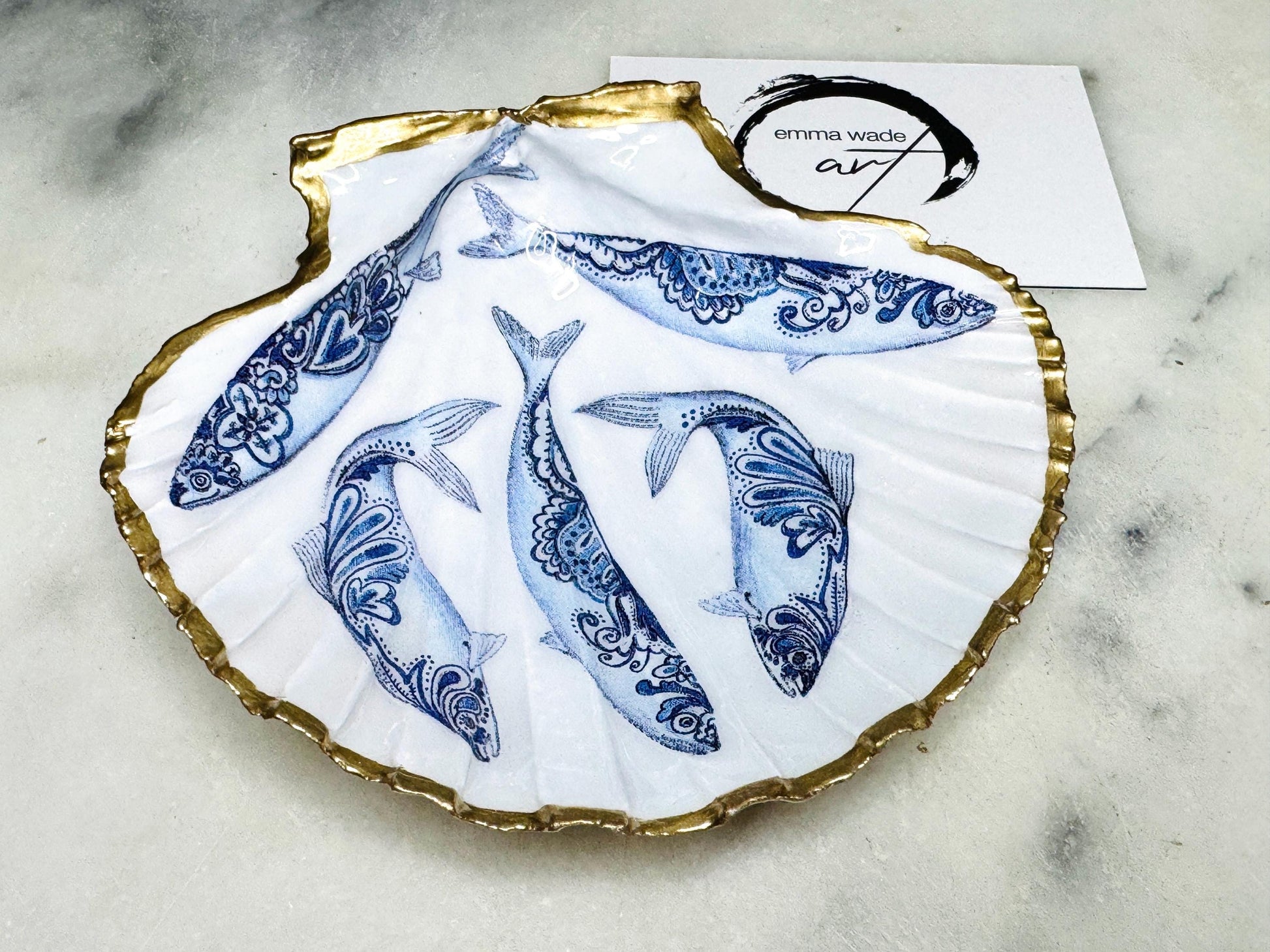 Handmade Dorset scallop shell trinket dishes featuring blue and white paisley fish illustrations with hand-painted gold leaf edging and glossy crystal clear resin. Coastal Scandinavian inspired jewellery dishes styled on a soft neutral background with elegant nautical fish artwork and luxurious gilded detailing.
