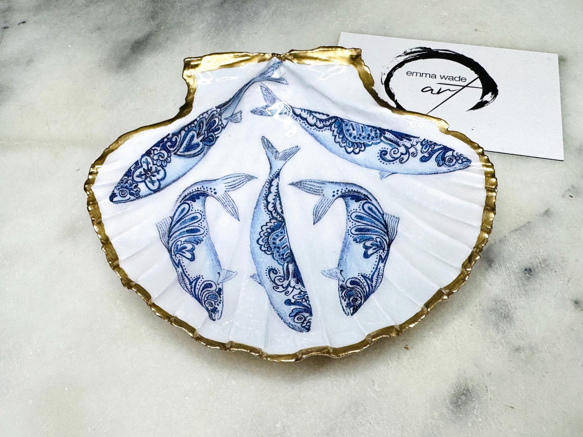 Handmade Dorset scallop shell trinket dishes featuring blue and white paisley fish illustrations with hand-painted gold leaf edging and glossy crystal clear resin. Coastal Scandinavian inspired jewellery dishes styled on a soft neutral background with elegant nautical fish artwork and luxurious gilded detailing.