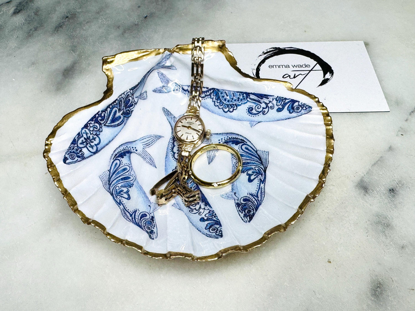 Handmade Dorset scallop shell trinket dishes featuring blue and white paisley fish illustrations with hand-painted gold leaf edging and glossy crystal clear resin. Coastal Scandinavian inspired jewellery dishes styled on a soft neutral background with elegant nautical fish artwork and luxurious gilded detailing.