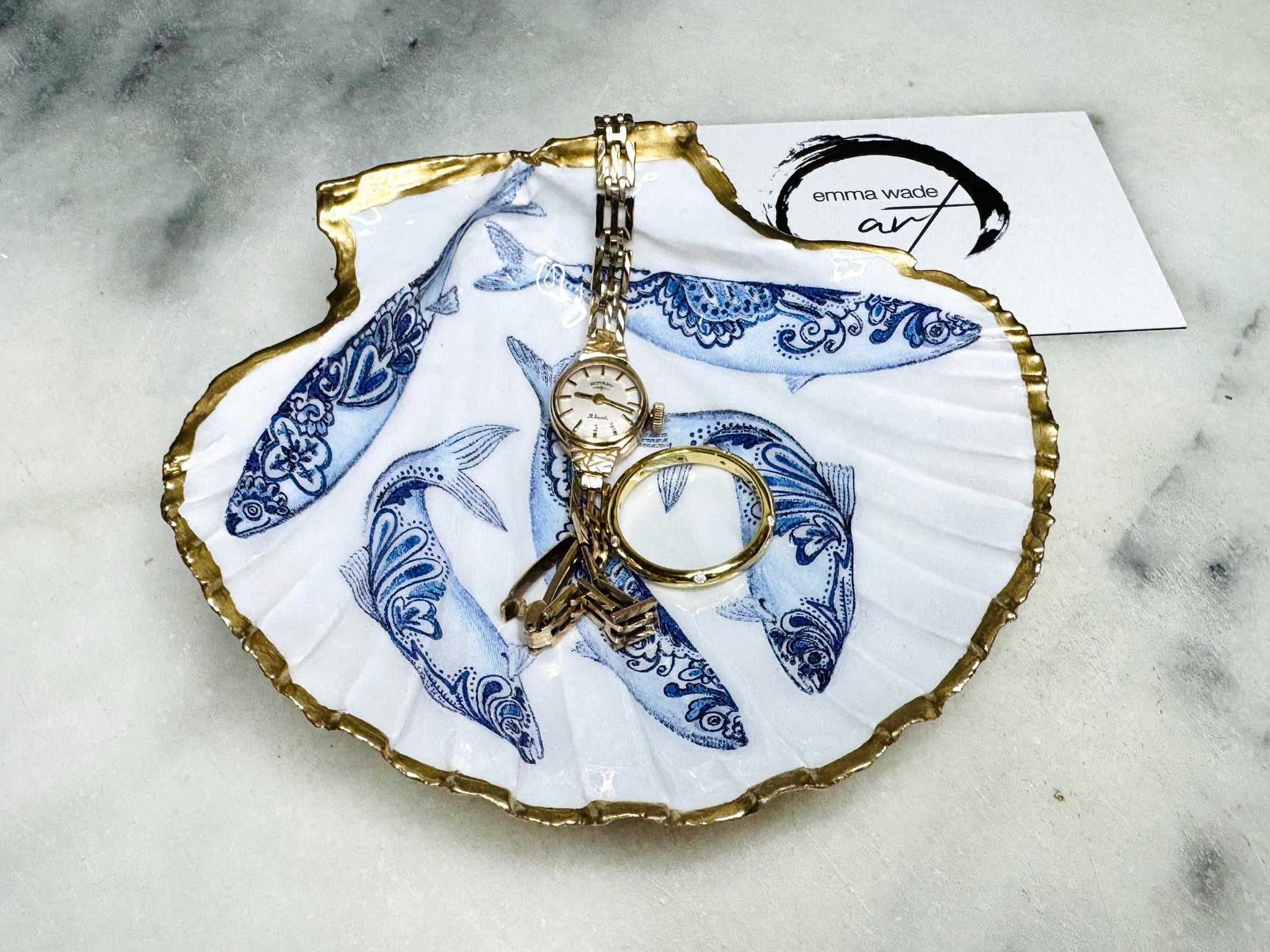 Handmade Dorset scallop shell trinket dishes featuring blue and white paisley fish illustrations with hand-painted gold leaf edging and glossy crystal clear resin. Coastal Scandinavian inspired jewellery dishes styled on a soft neutral background with elegant nautical fish artwork and luxurious gilded detailing.