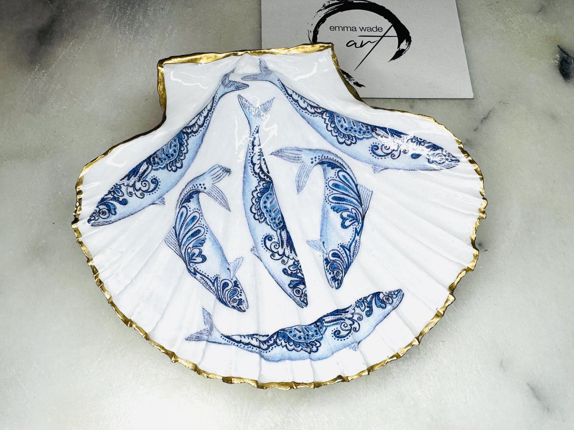 Handmade Dorset scallop shell trinket dishes featuring blue and white paisley fish illustrations with hand-painted gold leaf edging and glossy crystal clear resin. Coastal Scandinavian inspired jewellery dishes styled on a soft neutral background with elegant nautical fish artwork and luxurious gilded detailing.