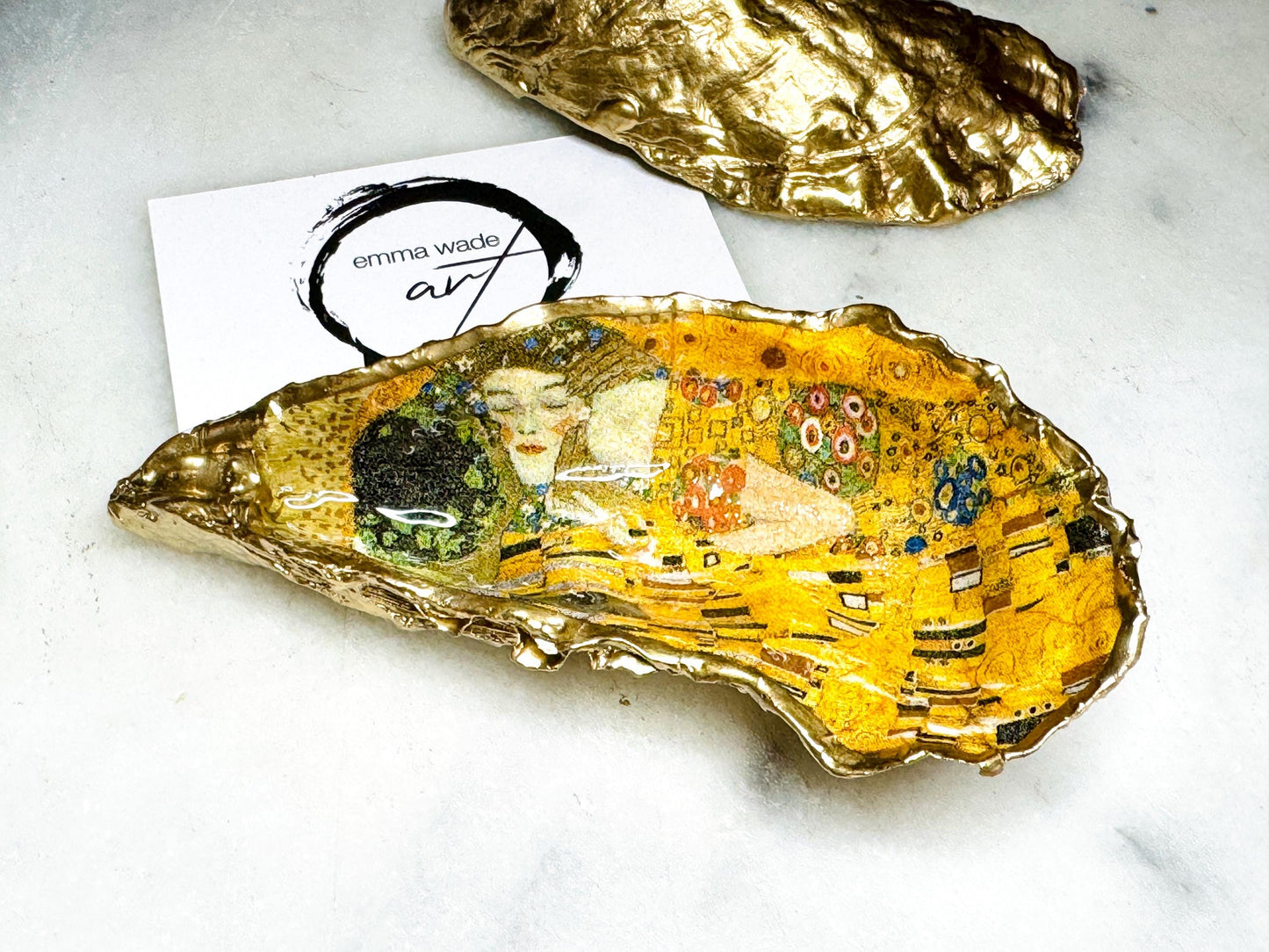 Handcrafted Dorset oyster shell trinket dish inspired by Gustav Klimt’s The Kiss, featuring romantic gold and green artwork detail. Natural oyster shell edged in hand applied liquid gold leaf and sealed with high gloss resin. Luxury art inspired jewellery holder displayed with gold rings on marble background. Unique gift for her or art lover keepsake.
