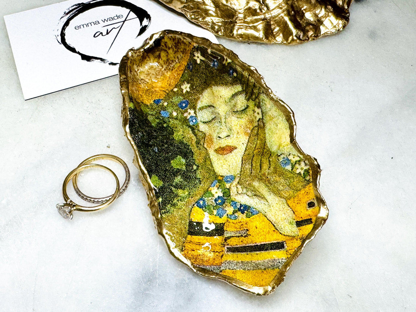 Handcrafted Dorset oyster shell trinket dish inspired by Gustav Klimt’s The Kiss, featuring romantic gold and green artwork detail. Natural oyster shell edged in hand applied liquid gold leaf and sealed with high gloss resin. Luxury art inspired jewellery holder displayed with gold rings on marble background. Unique gift for her or art lover keepsake.