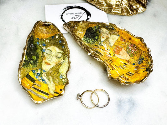 Handcrafted Dorset oyster shell trinket dish inspired by Gustav Klimt’s The Kiss, featuring romantic gold and green artwork detail. Natural oyster shell edged in hand applied liquid gold leaf and sealed with high gloss resin. Luxury art inspired jewellery holder displayed with gold rings on marble background. Unique gift for her or art lover keepsake.