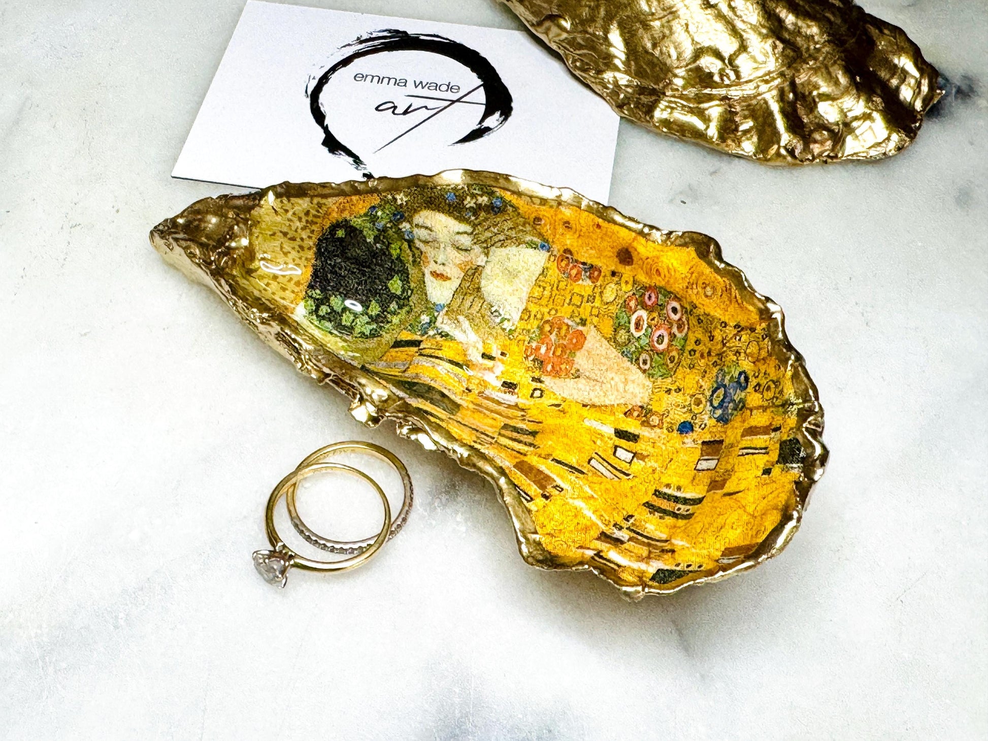 Handcrafted Dorset oyster shell trinket dish inspired by Gustav Klimt’s The Kiss, featuring romantic gold and green artwork detail. Natural oyster shell edged in hand applied liquid gold leaf and sealed with high gloss resin. Luxury art inspired jewellery holder displayed with gold rings on marble background. Unique gift for her or art lover keepsake.