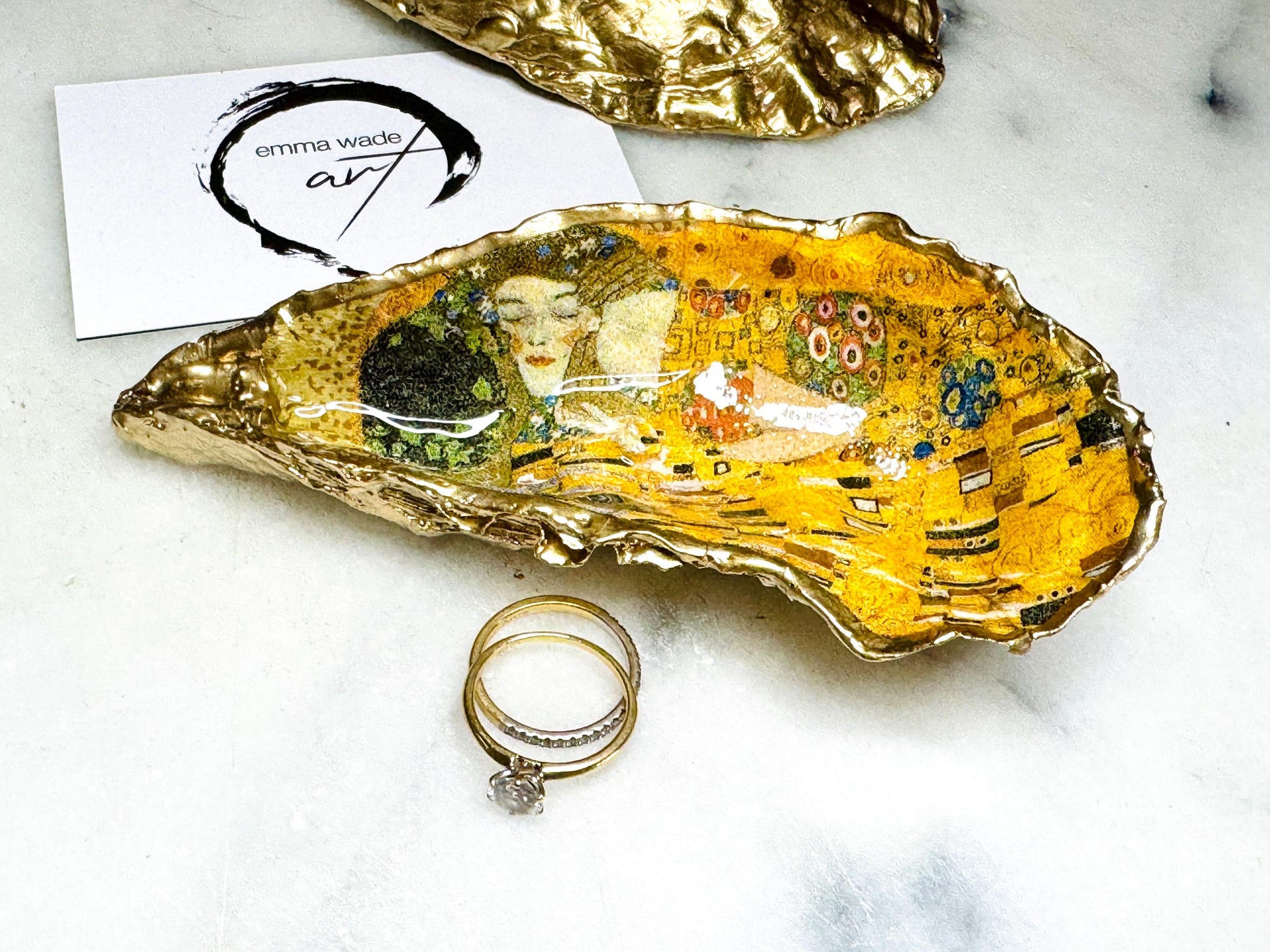 Handcrafted Dorset oyster shell trinket dish inspired by Gustav Klimt’s The Kiss, featuring romantic gold and green artwork detail. Natural oyster shell edged in hand applied liquid gold leaf and sealed with high gloss resin. Luxury art inspired jewellery holder displayed with gold rings on marble background. Unique gift for her or art lover keepsake.