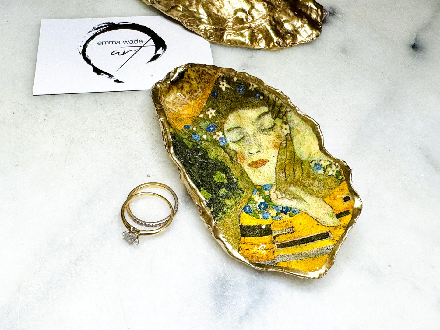 Handcrafted Dorset oyster shell trinket dish inspired by Gustav Klimt’s The Kiss, featuring romantic gold and green artwork detail. Natural oyster shell edged in hand applied liquid gold leaf and sealed with high gloss resin. Luxury art inspired jewellery holder displayed with gold rings on marble background. Unique gift for her or art lover keepsake.