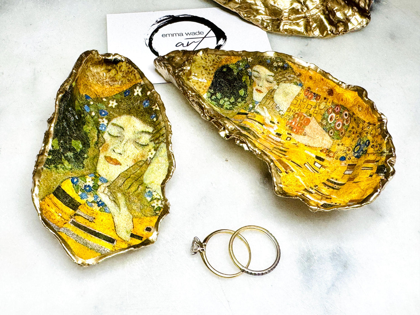 Handcrafted Dorset oyster shell trinket dish inspired by Gustav Klimt’s The Kiss, featuring romantic gold and green artwork detail. Natural oyster shell edged in hand applied liquid gold leaf and sealed with high gloss resin. Luxury art inspired jewellery holder displayed with gold rings on marble background. Unique gift for her or art lover keepsake.