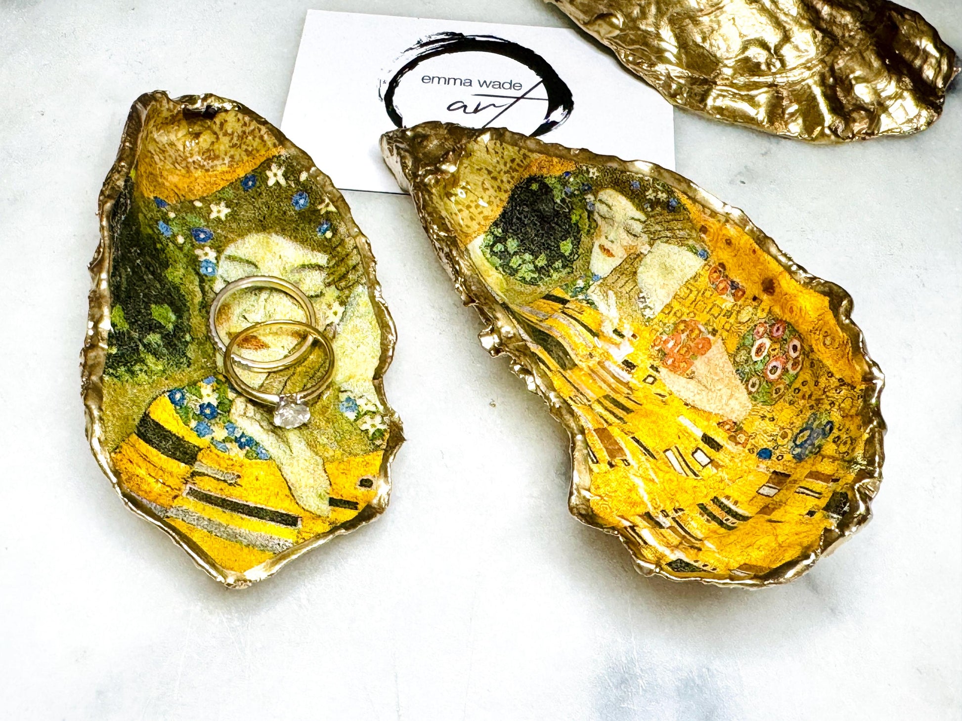 Handcrafted Dorset oyster shell trinket dish inspired by Gustav Klimt’s The Kiss, featuring romantic gold and green artwork detail. Natural oyster shell edged in hand applied liquid gold leaf and sealed with high gloss resin. Luxury art inspired jewellery holder displayed with gold rings on marble background. Unique gift for her or art lover keepsake.