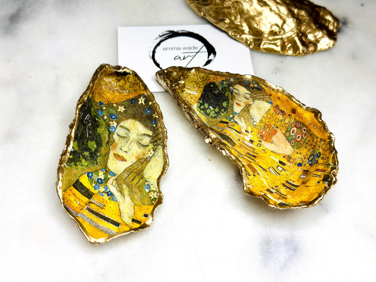 Handcrafted Dorset oyster shell trinket dish inspired by Gustav Klimt’s The Kiss, featuring romantic gold and green artwork detail. Natural oyster shell edged in hand applied liquid gold leaf and sealed with high gloss resin. Luxury art inspired jewellery holder displayed with gold rings on marble background. Unique gift for her or art lover keepsake.